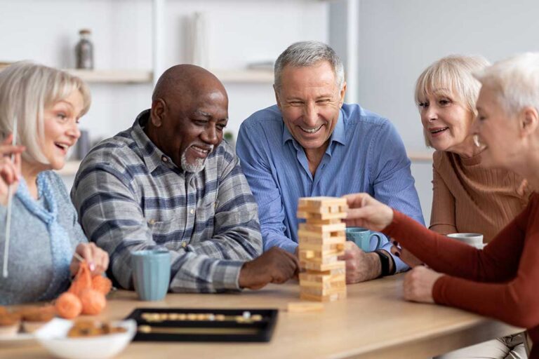 A senior woman sits on a bed surrounded by moving boxes while talking with family in a cozy, well-lit room.